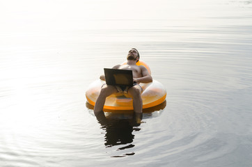 Fototapeta na wymiar A man with a laptop on an inflatable ring in the water at sunset, free space.