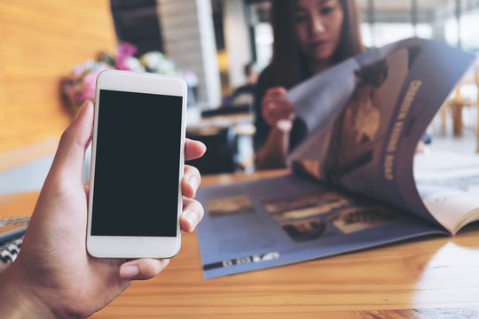 Mockup image of a man's hand holding white mobile phone with blank black screen in modern cafe and blur woman reading newspaper in background