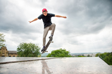 Long-haired skater-teenager in a T-shirt and a sneaker hat jumps an alley against a stormy sky