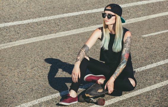 A beautiful, sexy hipster blonde with blue hair in tattoo sits on a longboard  with glasses and a cap on the asphalt on the street