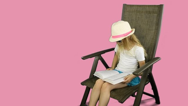 Young caucasian girl in white t-shirt and hat sitting on chair and reading book carefully.