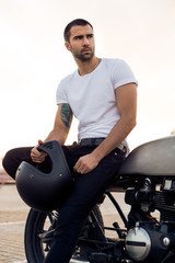 Close up of a handsome rider guy in white blank t-shirt look to the side near cafe racer motorbike at sunset. Vintage bike custom made in garage. Brutal urban lifestyle. Outdoor portrait. - 165006166