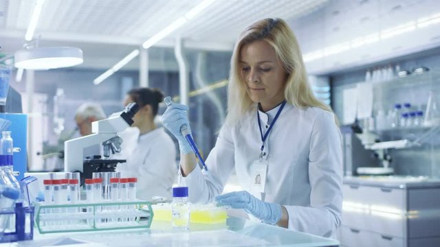 Female Research Scientist Uses Micropipette Filling Test Tubes in a Big Modern Laboratory. In the Background Scientists are Working. Shot on RED EPIC-W 8K Helium Cinema Camera.