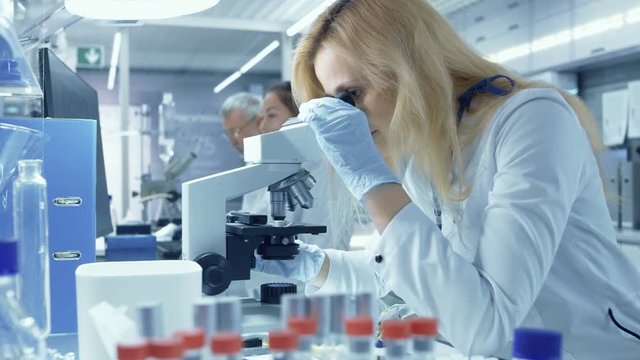 Female Research Scientist Looks at Biological Samples Under Microscope. She and Her Colleagues Work in a Big Modern Laboratory. Shot on RED EPIC-W 8K Helium Cinema Camera.