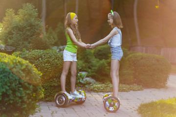 Happy girls riding on hover boards or gyroscooters outdoors at sunset in summer. Active life concept - obrazy, fototapety, plakaty