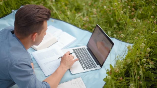 Young Handsome Man is Working on Laptop Lying on a Blanket in the Park in the Summer