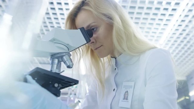 Low Angle Shot of Medical Research Scientists putting Slides in Place and Looking at Samples Under Microscope. She Works in a Bright Modern Laboratory. Shot on RED EPIC-W 8K Helium Cinema Camera.