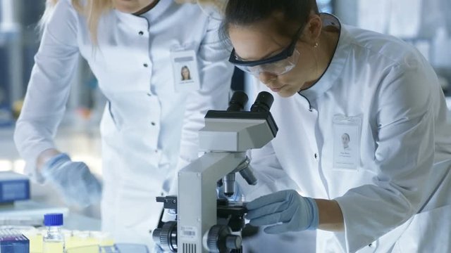 Medical Research Scientist Drops Sample on Slide and Her Colleague Examines it Under Microscope. They Work in a Modern Laboratory. Shot on RED EPIC-W 8K Helium Cinema Camera.