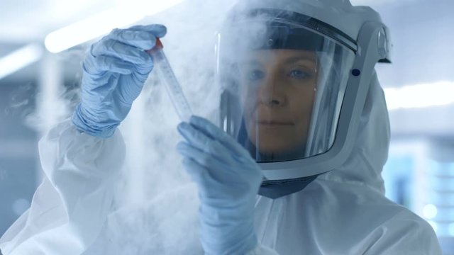 Medical Virology Research Scientist in a Hazmat Suit with Mask, She Inspects Test Tube with Isolated Virus String from Refrigerator Box. She Works in a Sterile High Tech Laboratory.