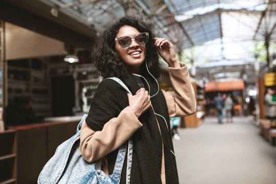 Cool African American girl wearing sunglasses and earphones. Smiling girl with dark curly hair looking in camera on street. Nice girl in coat,scarf and bag on her shoulder walking around street
