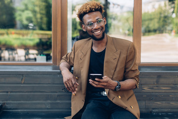 A portrait of African American man in jacket and glasses with smartphone sitting near the windows