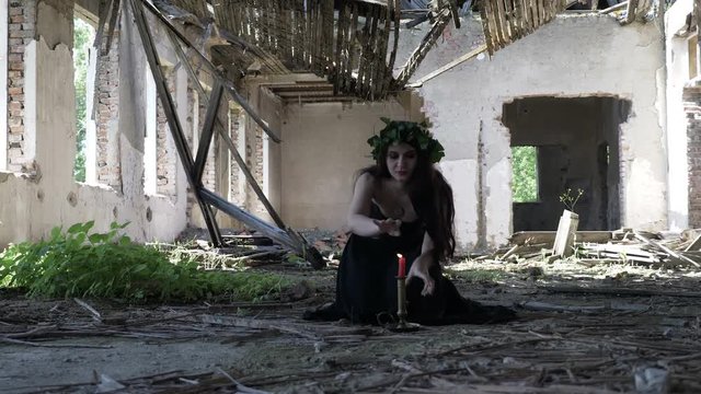 Beautiful witch conjuring and invoking maleficent spirits with incantation and candles in an abandoned ruined mansion