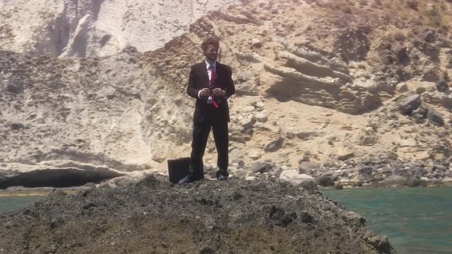 Man in suit stranded on a deserted rock in the middle of the ocean looking around with binoculars