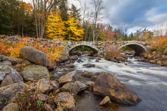 Stone Bridge Over A Flowing River In New Hampshire With Fall Foliage