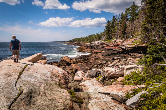 Fototapeta   A man walks on large boulder surrounded by rocky shoreline and forest during a hike on a beautiful summer day