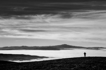 A man in a mountains scenery, with fog filling a valley and a big, deep sky