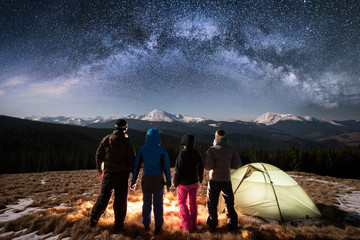 Silhouette of four people standing together beside camp and tent under beautiful night sky full of stars and milky way. On the background snow-covered mountains. Rear view. Long exposure