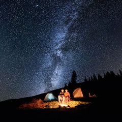 Night camping in the mountains. Couple tourists have a rest at a campfire near two tents under beautiful night sky full of stars and milky way. Long exposure