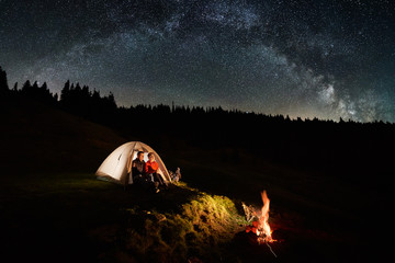 Night camping in the mountains. Couple tourists have a rest in the illuminated tent near campfire under beautiful night sky full of stars and milky way. Low light © anatoliy_gleb