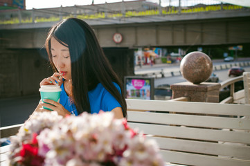 Cute asian young woman in summer cafe outdoors. girl In a blue dress, with long hair in simple light cozy interior of restaurant Urban style, drinking coffee drink, sitting at table with flowers