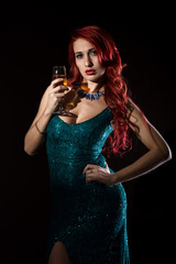 Naklejka premium Young redhead sexy woman in blue elegant dress holding wineglass with champagne and poses in a dark.