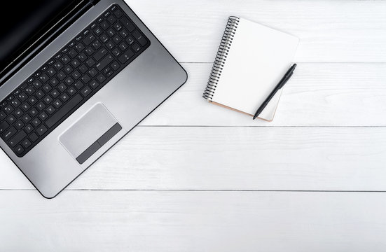 Wooden white office desk table with open blank laptop computer and empty white diary with pen, free space. Top view with copy space. Grey laptop on wooden background