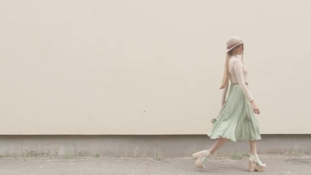 Model in a hat and a long skirt walks on the camera against the wall