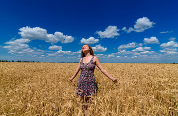 Portrait of beautiful young woman in dress walking in golden wheat field with cloudy blue sky background, free space. Liberty, peace of mind concept. Girl in spikes of ripe wheat field under blue sky 