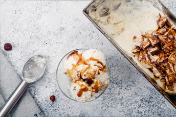 Caramel ice cream scooped in glass bowl from metallic container. Peanuts and chocolate used for decoration. Top view with copy space. Rustic textured background.