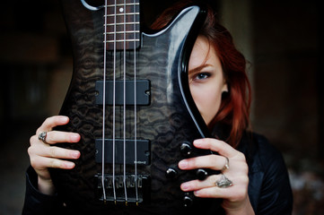 Red haired punk girl wear on black with bass guitar at abadoned place. Portrait of gothic woman musician. Close up face of blackness person with guitar riff.
