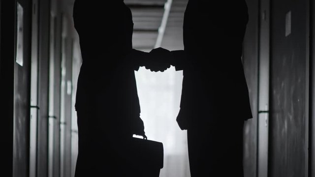 Silhouettes of unrecognizable businessman with briefcase and his business partner standing in dark hallway and exchanging handshake in agreement 