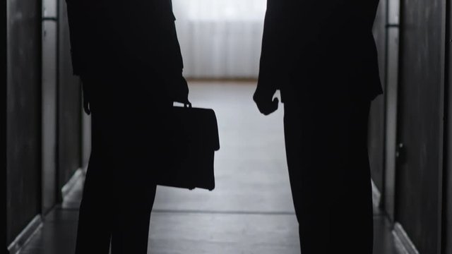 Tilt up of silhouettes of businessman holding briefcase and his colleague shaking hands in dark hallway, then walking away