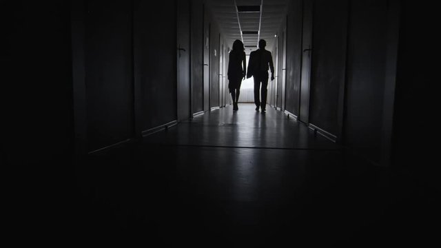 PAN of silhouettes of businesswoman holding briefcase and her male colleague walking along dark corridor of business centre 
