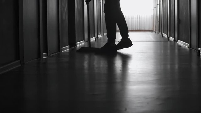 PAN with low-section of unrecognizable figure of male janitor sweeping floor in dark corridor