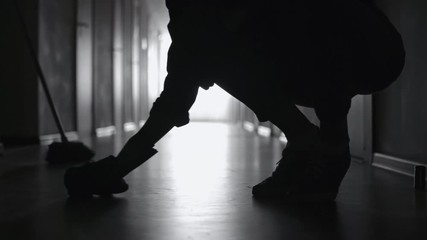 PAN with low-section of silhouette of legs of female janitor scrubbing floor in dark corridor
