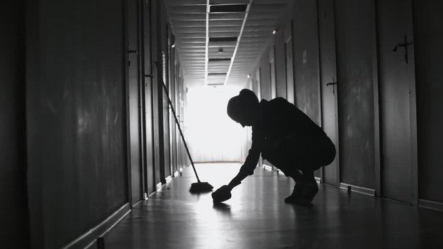PAN of silhouette of tired female janitor scrubbing floor and wiping sweat of her face in dark corridor 