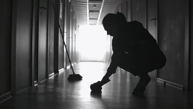 PAN with of silhouette of female janitor manually scrubbing floor in dark corridor, then wiping sweat of her face 