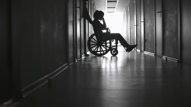 PAN with slow motion of silhouette of male healthcare worker pushing wheelchair with sad female patient out of room, then walking along dark hallway 