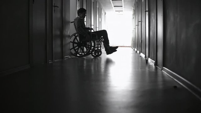 PAN with low angle of paraplegic young man riding wheelchair appearing out of hospital room, then heading along dark corridor towards bright light coming from window 