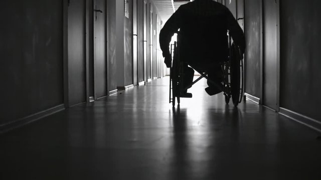 Pan with low angle of silhouette of disabled man in wheelchair riding along dark hospital corridor