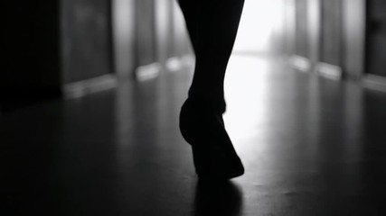 Tracking with low-section of silhouette of female legs in high heels shoes walking along dark hallway; black and white slow motion shot - Powered by Adobe