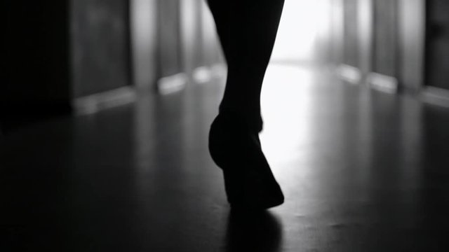 Tracking with low-section of silhouette of female legs in high heels shoes walking along dark hallway; black and white slow motion shot