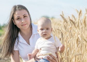 Young pretty mother with her adorable baby boy sitting in wheat field at summer day