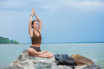 Fototapeta na wymiar Girl practicing yoga on the rocks against the blue sky and the azure sea. Woman raises her arms to the sky in namaste posture and sits in a lotus pose.