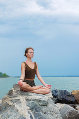 Girl practicing yoga on the rocks against the blue sky and the azure sea. Woman raises her arms to the sky in namaste posture and sits in a lotus pose.