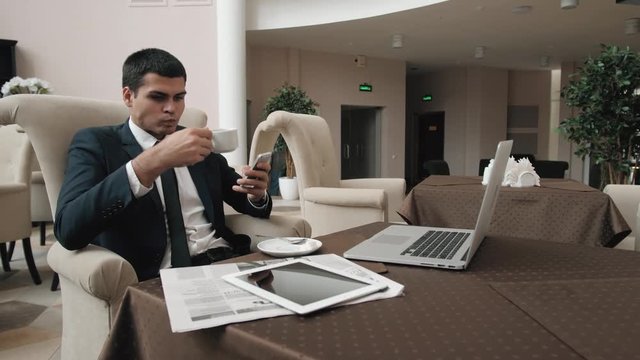Businessman using smart phone while coffee break in convention center, man in hotel lobby wait taxi to airport and work with phone and laptop