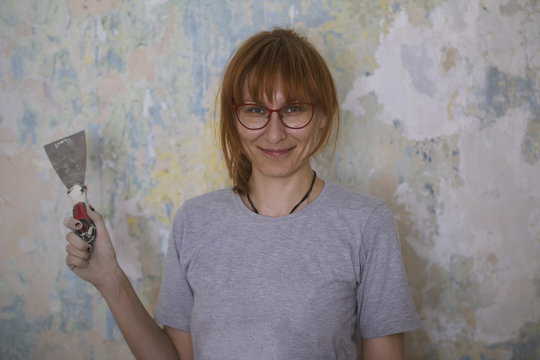 Home renovation - happy red hair woman with scraper near wall glues Wallpaper