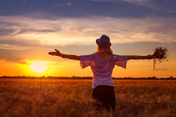 Background girl in a hat with a bouquet of wildflowers in one hand looks at the evening contras sunset standing on the field of ripe wheat and another hand showing peace - obrazy, fototapety, plakaty