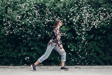 hipster woman in sunglasses walking on background of blooming bush with white flowers of spirea. boho girl outing in floral and denim modern clothes in summer city street. space for text