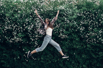 happy hipster woman in sunglasses having fun on background of blooming bush with white flowers of spirea. boho girl jumping and smiling in modern clothes, emotional moment. space for text